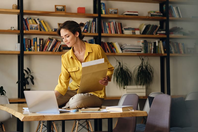 Young attractive smiling woman in shirt sitting on desk with papers dreamily working on laptop