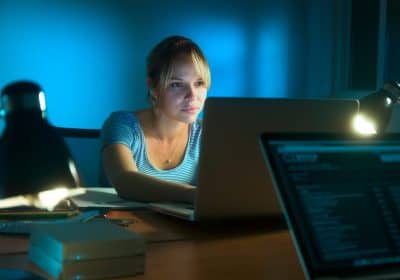 Beautiful woman working late at night in office, surfing the web and writing post on social network with laptop computer. The girl is concentrated and serious